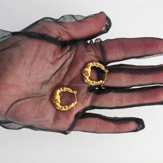 Gold hoop earrings held in a hand wearing a black sheer glove on a white background