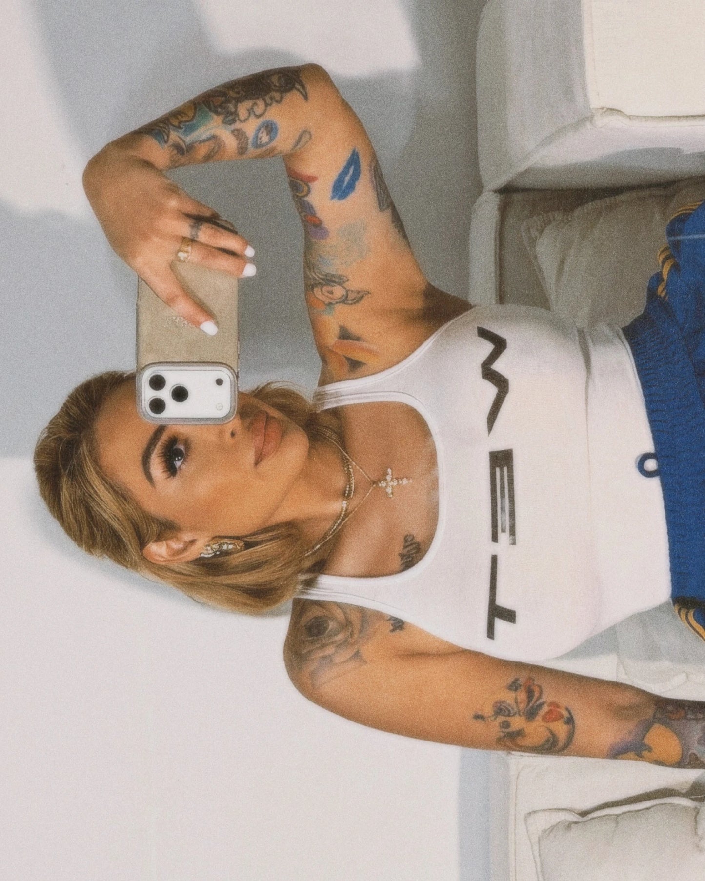 WOMEN with tattoos taking a selfie in a white tank top and blue jeans.